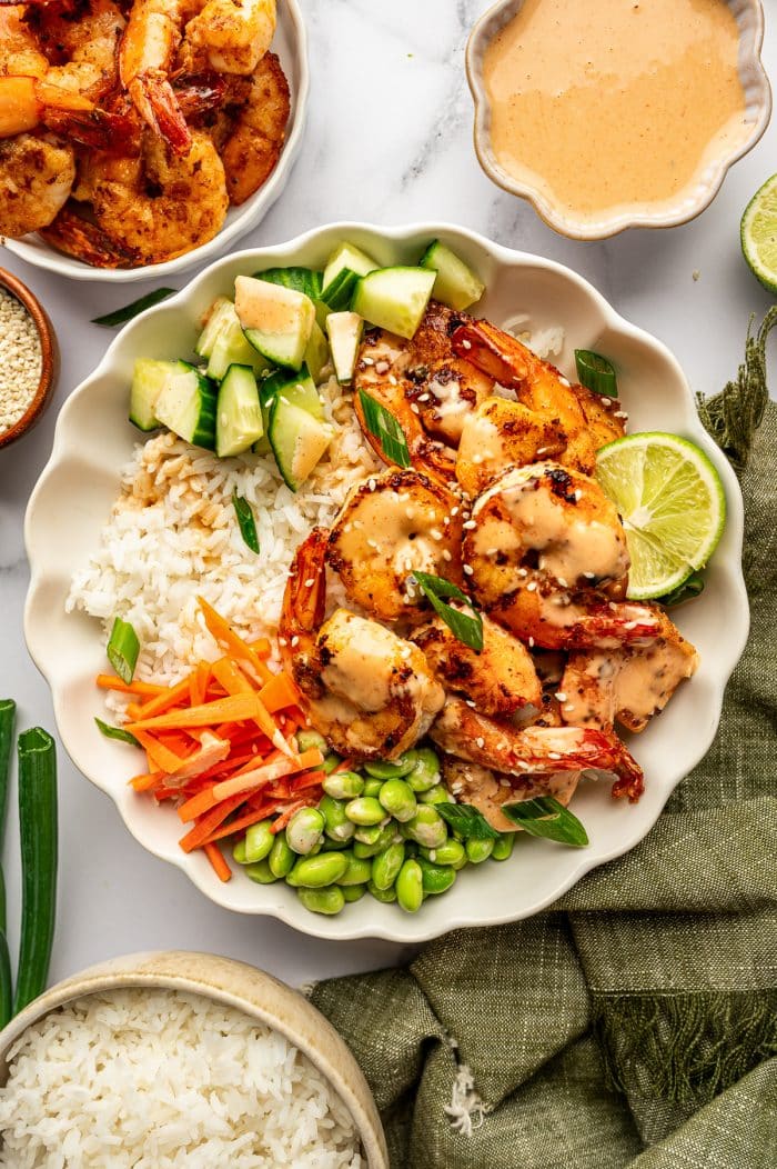 Overhead view of a complete shrimp rice bowl in a large ivory scalloped bowl filled with white jasmine rice, golden pan-seared shrimp, diced cucumber, matchstick carrots, and bright green edamame. The shrimp are drizzled with creamy orange spicy mayo and garnished with sliced green onions, white sesame seeds, and a lime wedge. A white bowl of extra shrimp, an ivory bowl of spicy mayo, a small brown bowl of sesame seeds, and a beige bowl of rice surround the main bowl on a white marble surface with an olive-green fringed cloth.