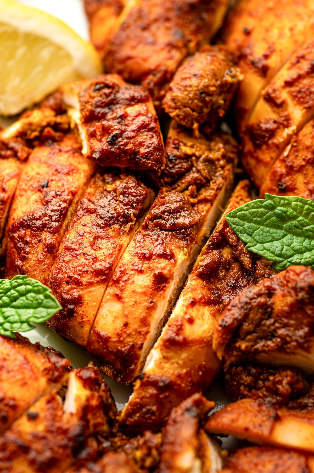 Extreme close-up of sliced chicken shawarma showing the juicy pale interior and deeply browned orange-red crust, garnished with bright green mint leaves and a blurred yellow lemon wedge.
