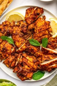 Sliced orange-red chicken shawarma arranged across a round white plate with yellow lemon wedges and fresh green mint leaves, with tan pita bread and a white bowl of tzatziki partially visible around the platter.