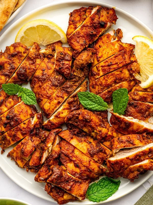 Sliced orange-red chicken shawarma arranged across a round white plate with yellow lemon wedges and fresh green mint leaves, with tan pita bread and a white bowl of tzatziki partially visible around the platter.
