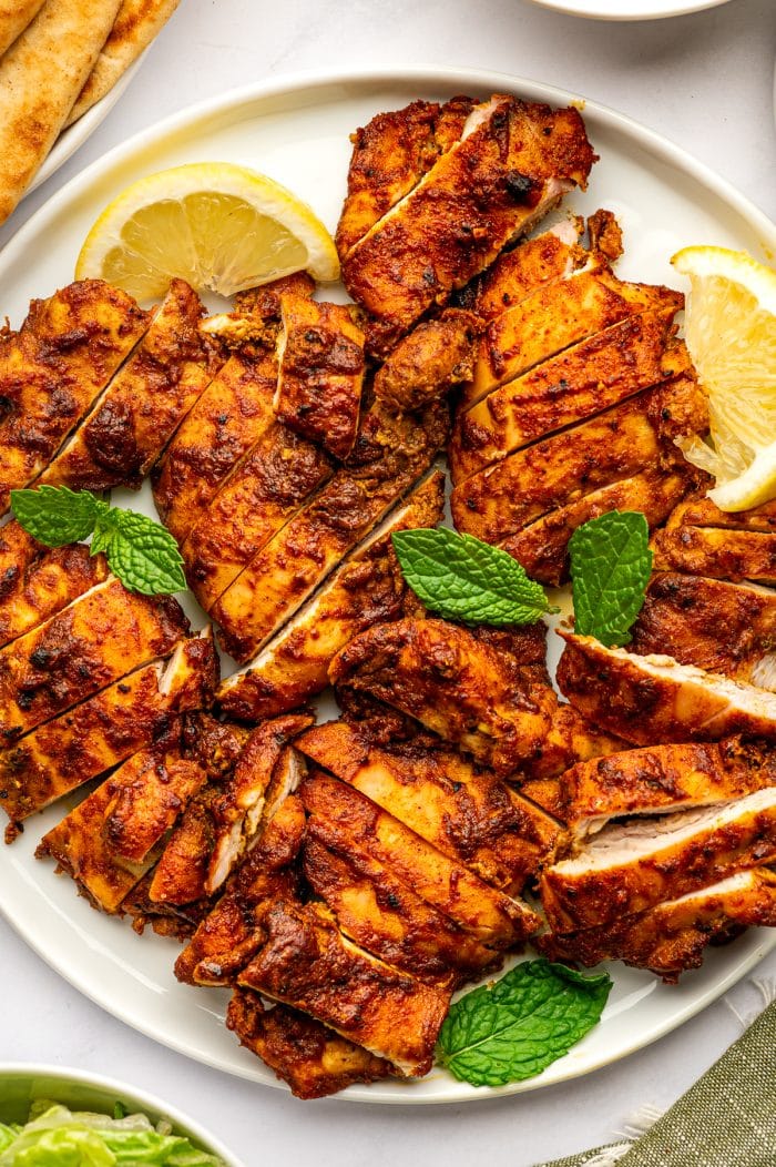 Sliced orange-red chicken shawarma arranged across a round white plate with yellow lemon wedges and fresh green mint leaves, with tan pita bread and a white bowl of tzatziki partially visible around the platter.
