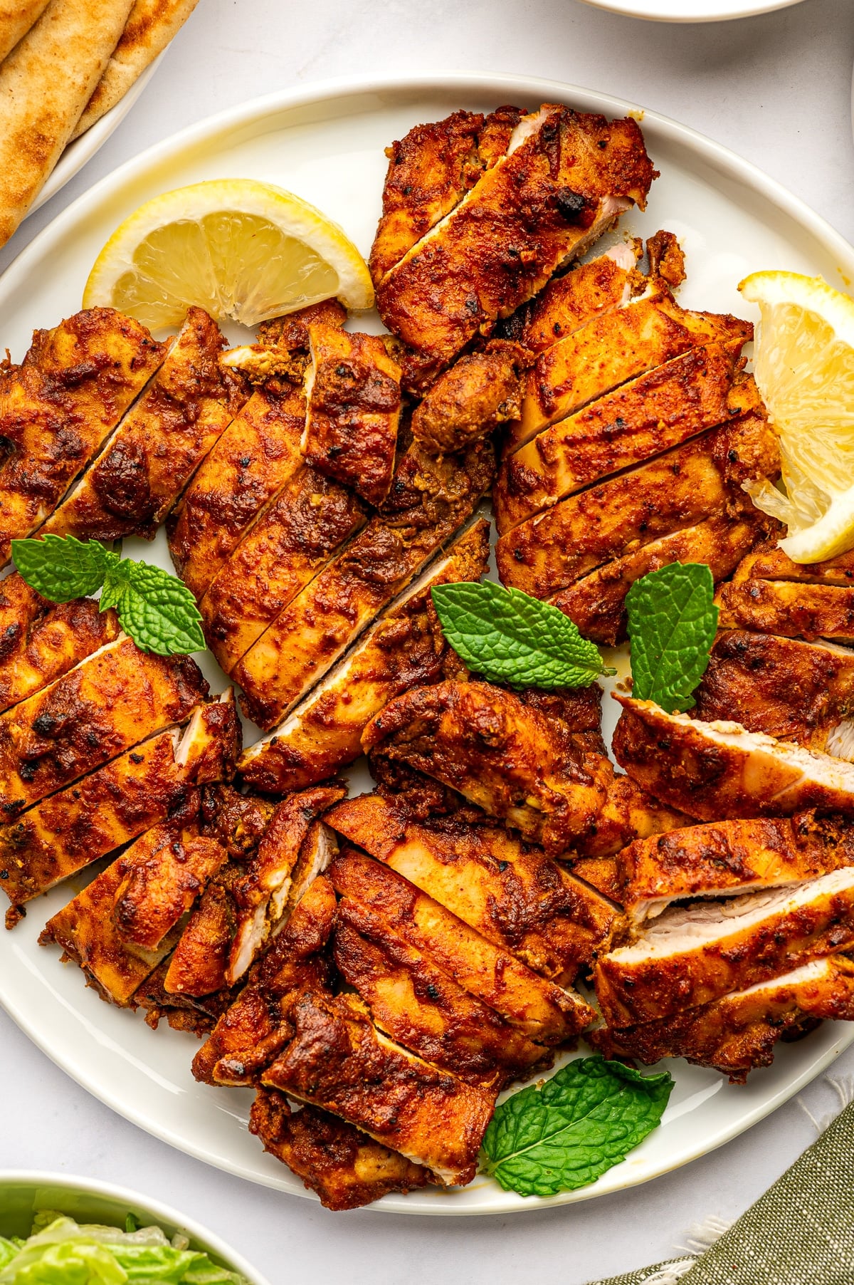 Sliced orange-red chicken shawarma arranged across a round white plate with yellow lemon wedges and fresh green mint leaves, with tan pita bread and a white bowl of tzatziki partially visible around the platter.