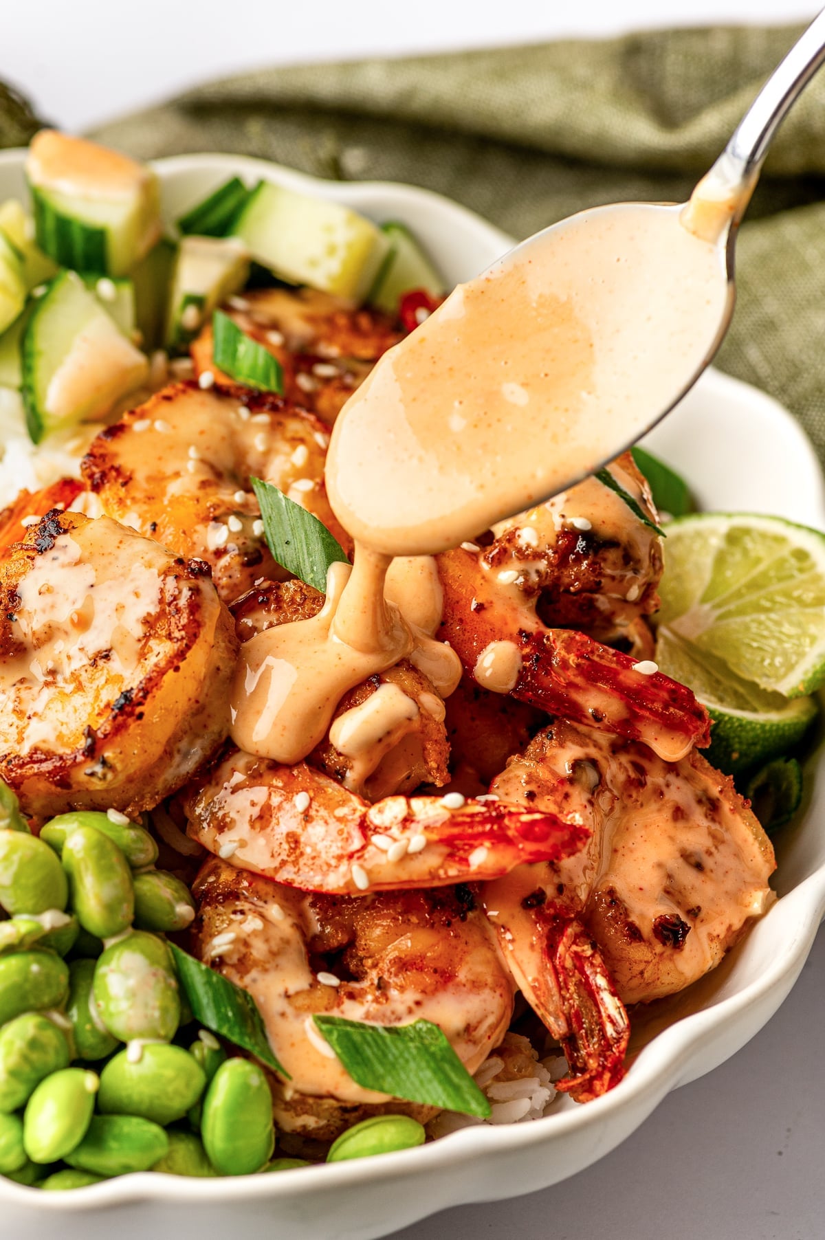 Close-up of a silver spoon pouring thick, pale orange spicy mayo over golden pan-seared shrimp in an ivory scalloped rice bowl. The bowl also contains bright green edamame, diced cucumber, white rice, sliced green onions, white sesame seeds, and a lime wedge, with an olive-green cloth in the background.