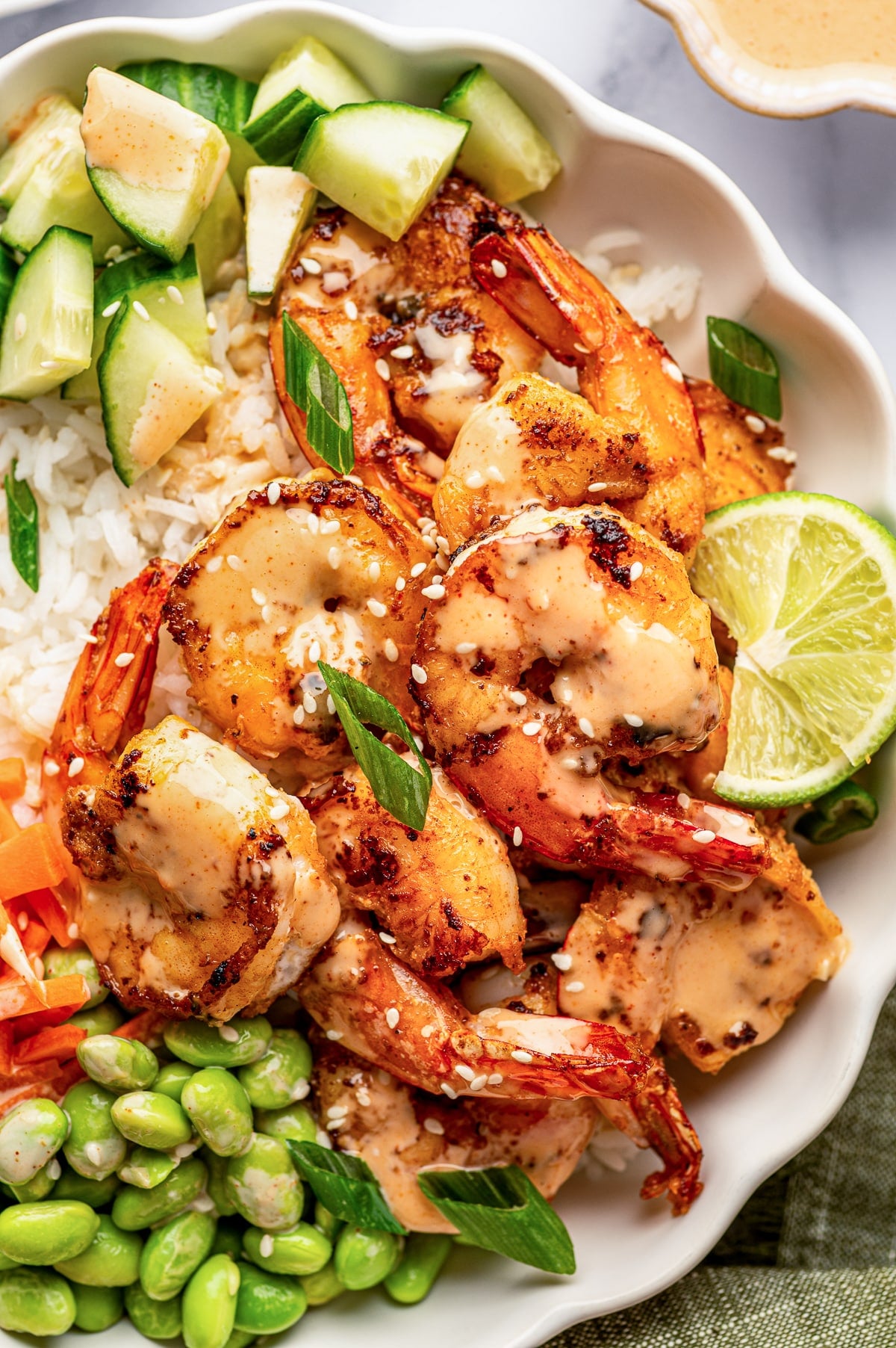 Close overhead view of an ivory scalloped bowl filled with white jasmine rice, browned shrimp with tails on, diced cucumber, matchstick carrots, and green edamame. Creamy orange spicy mayo is drizzled over the shrimp, which are garnished with white sesame seeds and sliced green onions, with a fresh lime wedge tucked along the side. Part of a small bowl of extra sauce appears at the upper right.