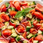Vertical Pinterest image showing a close-up of tomato cucumber salad in a white bowl with red tomato wedges, chopped green cucumber, thinly sliced red onion, parsley, black pepper, and a fresh mint garnish. A white text box reads "Tomato Cucumber Salad" and "quick + easy" in black text with a yellow divider line, and "THEFORKEDSPOON.COM" appears along the bottom.