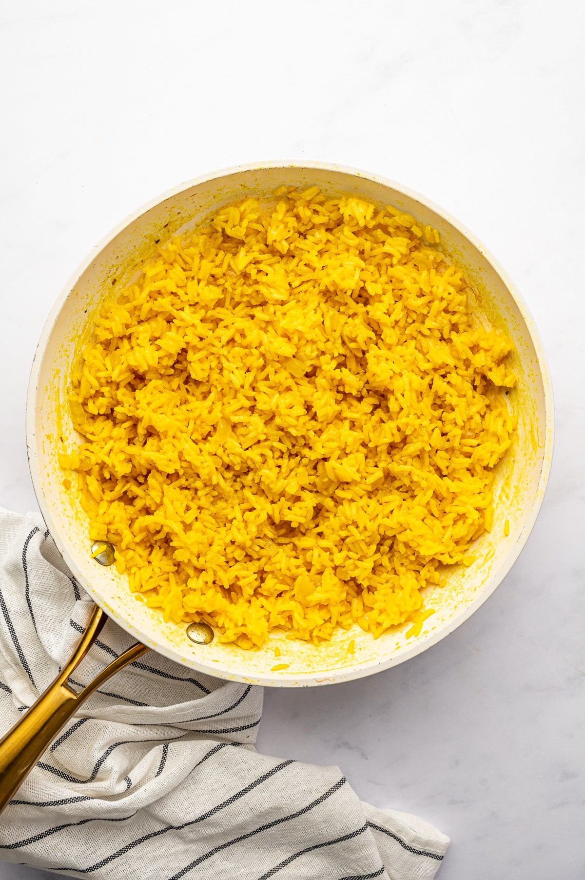 Fluffy golden turmeric yellow rice in a large white skillet with gold handles, photographed from overhead beside a white kitchen towel with thin black stripes.