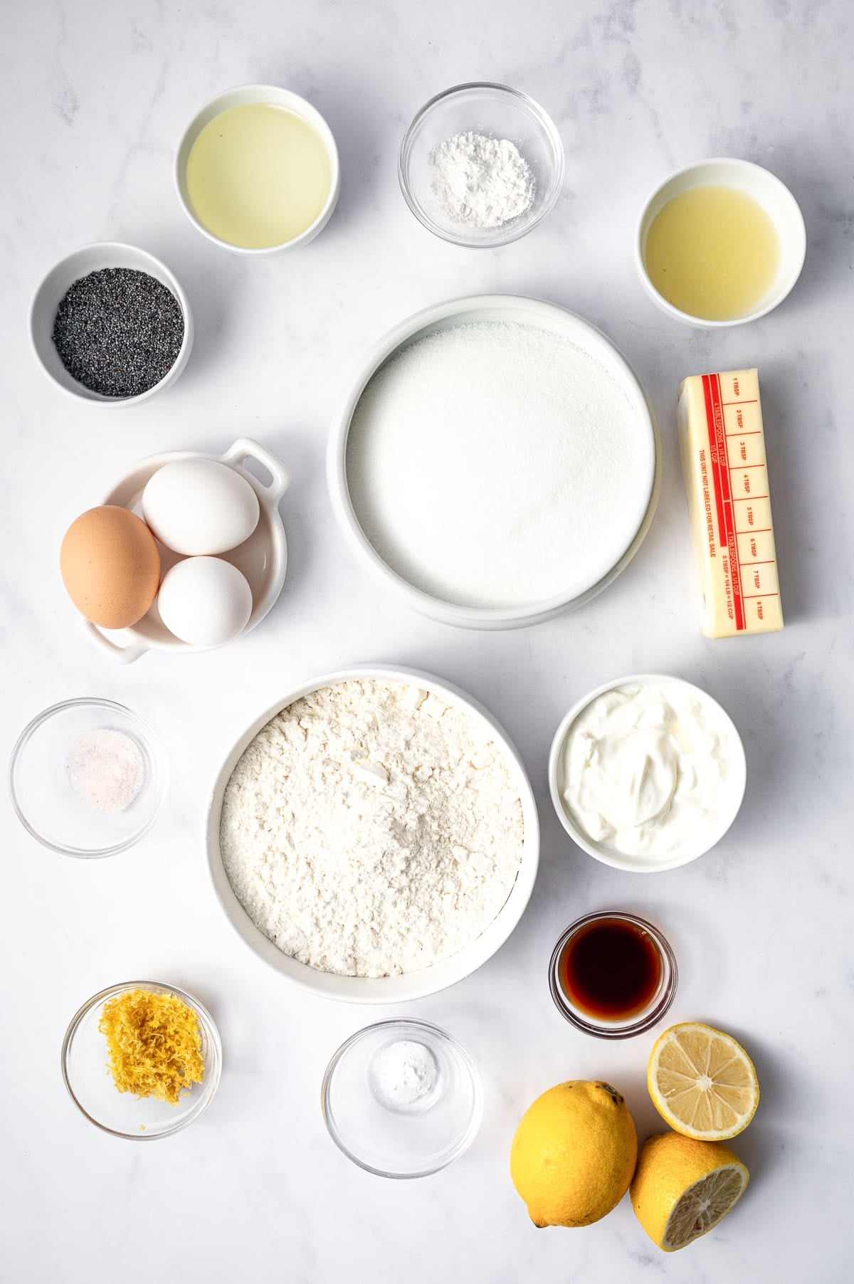 Overhead flat lay of lemon poppy seed bread ingredients arranged in white ceramic and clear glass bowls on a white marble surface.