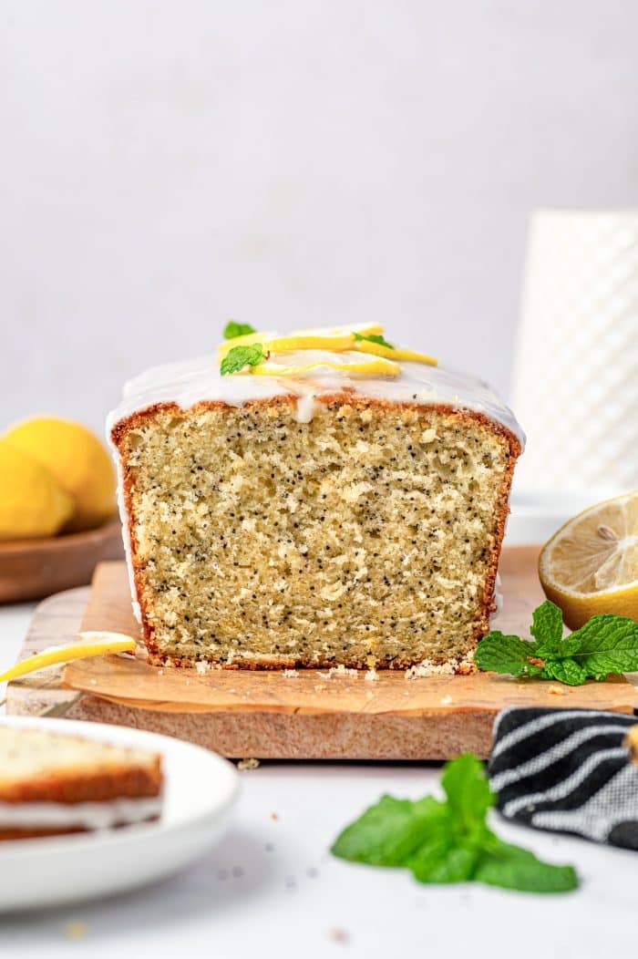 Front view of a sliced lemon poppy seed loaf on a rectangular wooden cutting board, showing the pale yellow, moist crumb evenly speckled with black poppy seeds and topped with white lemon glaze, thin lemon slices, and fresh green mint. A halved lemon, whole lemons, mint leaves, a black-and-white striped towel, and a white plate with slices of bread are arranged around the loaf.