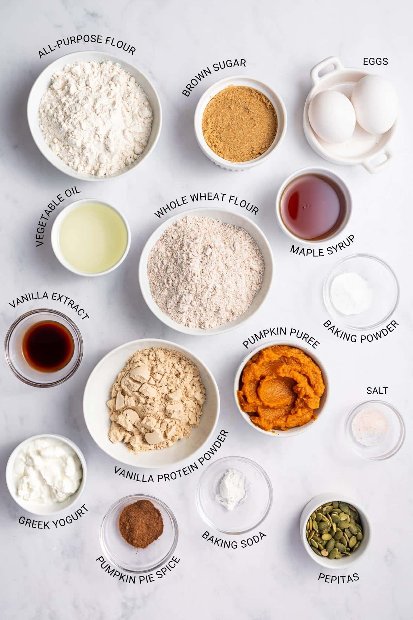 Overhead ingredient layout for protein pumpkin muffins on a white marble surface, with ingredients arranged in white bowls and individually labeled with black text. Labels read ALL-PURPOSE FLOUR, BROWN SUGAR, EGGS, VEGETABLE OIL, WHOLE WHEAT FLOUR, MAPLE SYRUP, VANILLA EXTRACT, BAKING POWDER, PUMPKIN PUREE, SALT, VANILLA PROTEIN POWDER, GREEK YOGURT, BAKING SODA, CINNAMON, and PEPITAS.