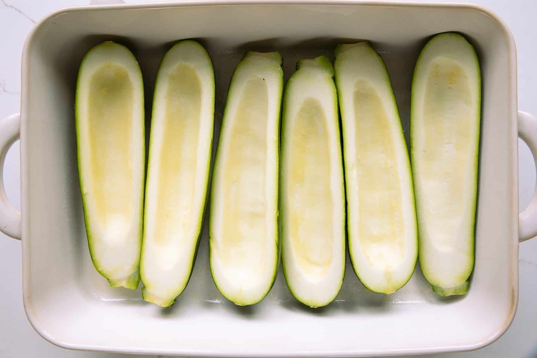 White baking dish filled with six zucchini halved with the seeds scraped out.