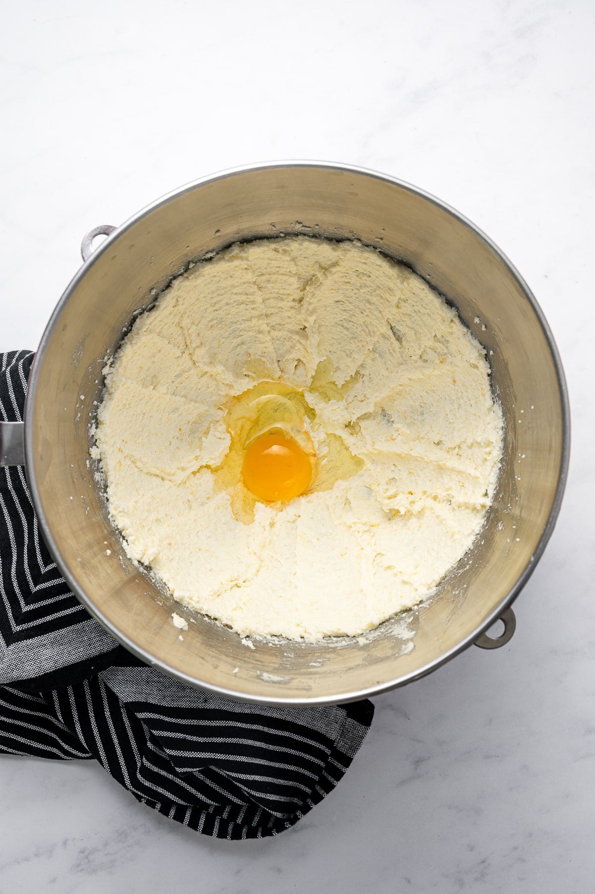 Overhead view of a raw egg added to pale, fluffy creamed butter, sugar, and lemon zest in a large stainless steel stand-mixer bowl. A black-and-white striped kitchen towel rests beside the silver bowl on a white marble surface.