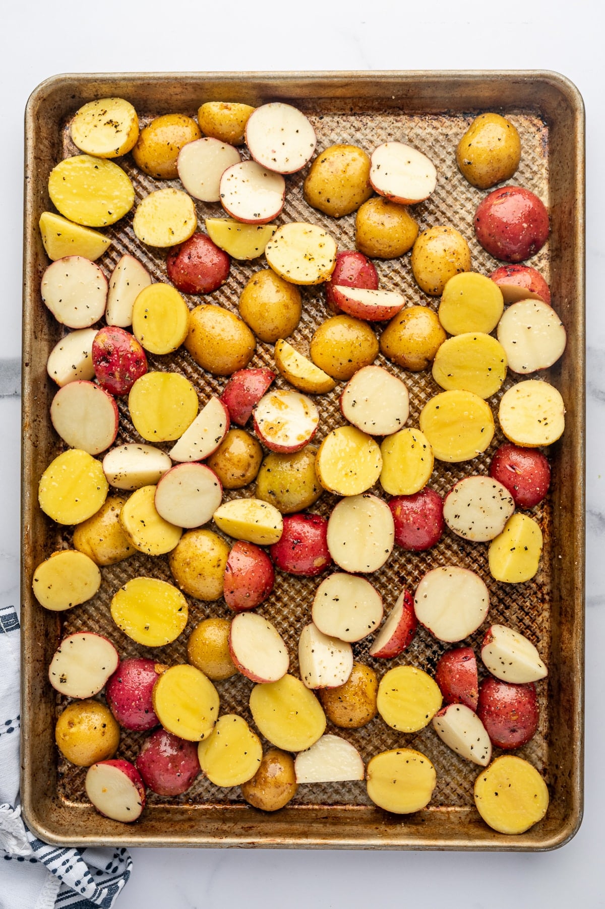 Seasoned baby Yukon Gold and red potatoes spread in a single layer across a large well-used metal rimmed baking sheet, with many of the potatoes positioned cut-side down and space between the pieces.