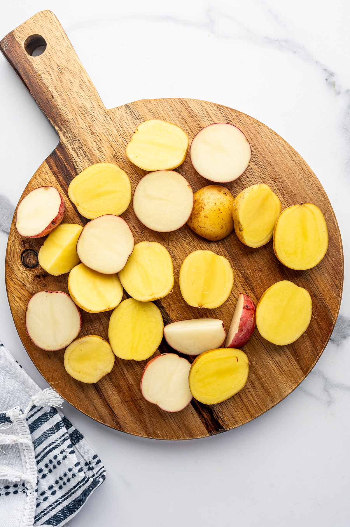 Halved baby Yukon Gold and red potatoes arranged cut-side up and down on a round wooden cutting board on a white marble surface, with a blue-and-white striped kitchen towel in the lower left corner.