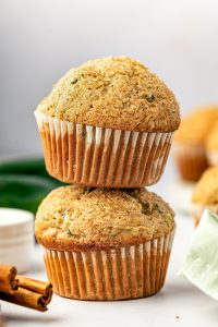 Two tall golden zucchini muffins stacked on a white surface, with white paper liners, coarse sparkling sugar on the domed tops, and green zucchini flecks throughout. Whole dark green zucchini, cinnamon sticks, and a pale green plaid napkin appear softly in the background.