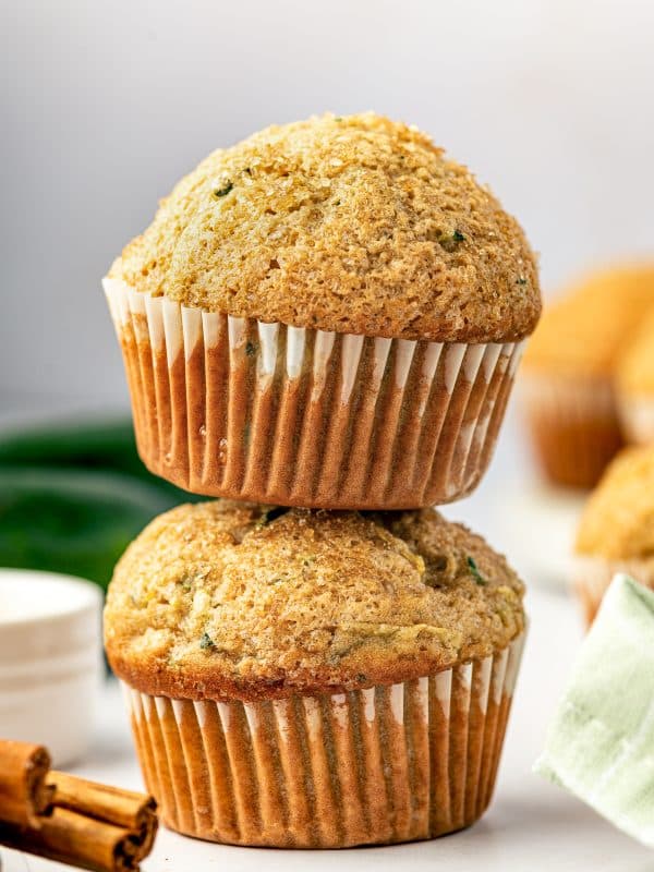 Two tall golden zucchini muffins stacked on a white surface, with white paper liners, coarse sparkling sugar on the domed tops, and green zucchini flecks throughout. Whole dark green zucchini, cinnamon sticks, and a pale green plaid napkin appear softly in the background.