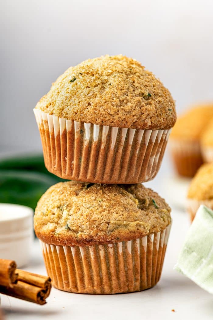 Two tall golden zucchini muffins stacked on a white surface, with white paper liners, coarse sparkling sugar on the domed tops, and green zucchini flecks throughout. Whole dark green zucchini, cinnamon sticks, and a pale green plaid napkin appear softly in the background.