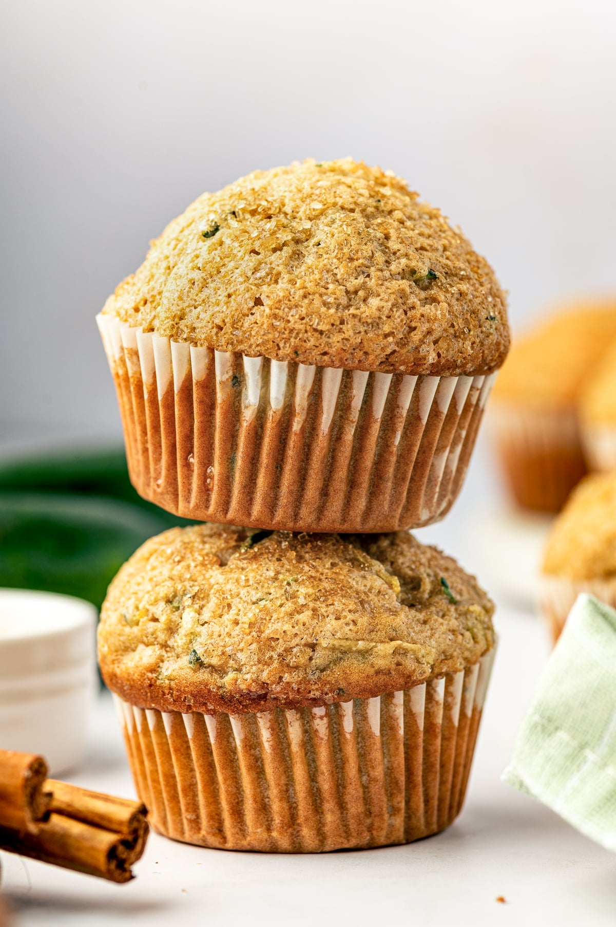 Two tall golden zucchini muffins stacked on a white surface, with white paper liners, coarse sparkling sugar on the domed tops, and green zucchini flecks throughout. Whole dark green zucchini, cinnamon sticks, and a pale green plaid napkin appear softly in the background.