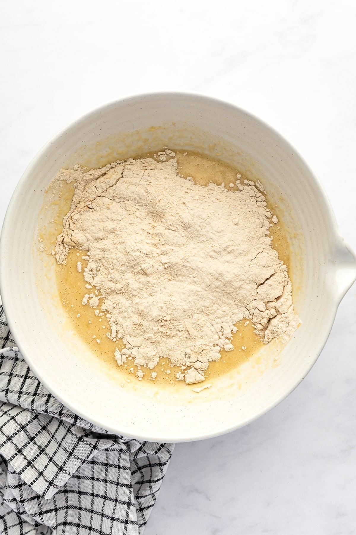 A mound of pale flour and vanilla protein powder mixture added to banana muffin batter in a large cream-colored mixing bowl with a handle, beside a black-and-white checkered towel.