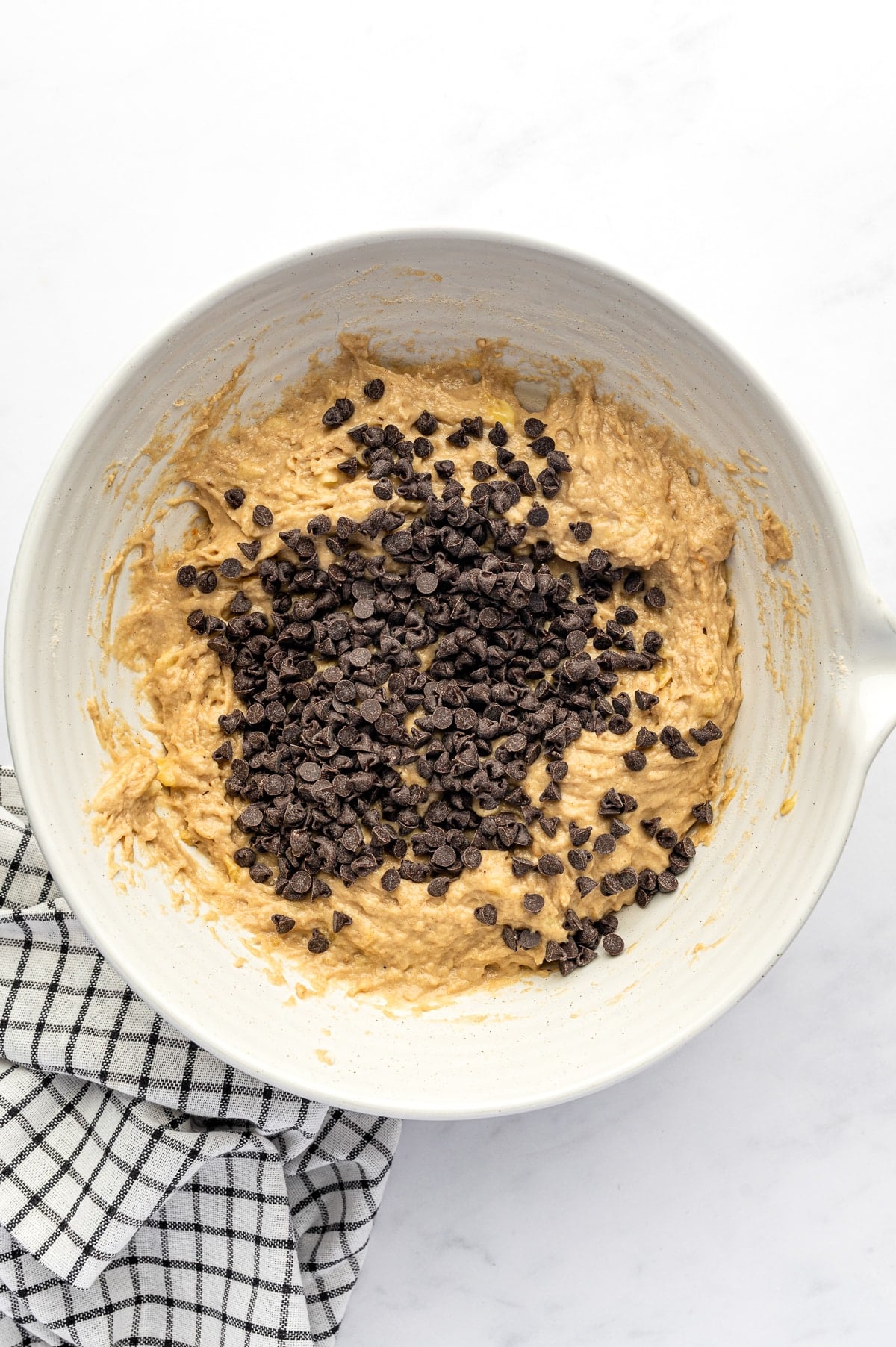 Thick tan banana protein muffin batter topped with mini semisweet chocolate chips in a large cream-colored mixing bowl, set beside a black-and-white checkered towel.