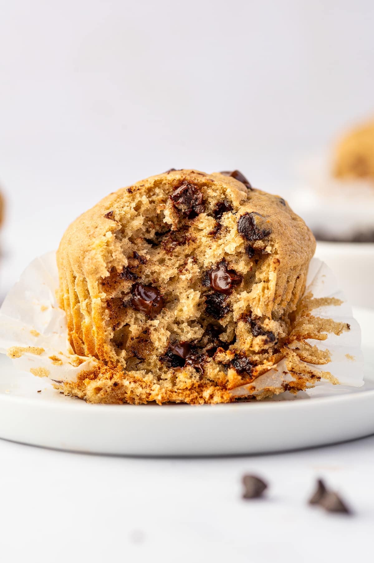 Banana protein muffin torn open to reveal a soft, moist golden crumb with melted mini chocolate chips, resting in an opened white paper liner on a white plate.