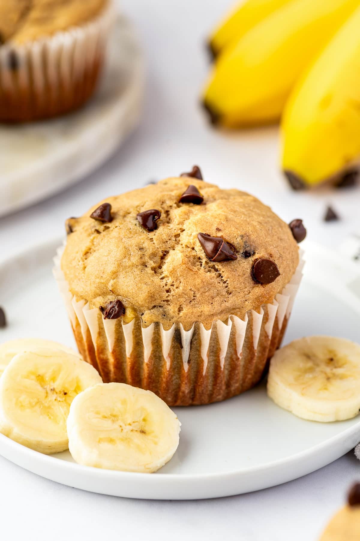 Golden banana protein muffin topped with mini chocolate chips in a white paper liner on a white plate, surrounded by fresh banana slices with whole yellow bananas softly blurred in the background.