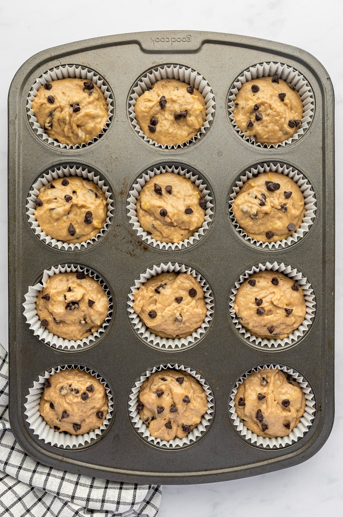 Twelve white paper muffin liners filled with tan banana protein muffin batter and topped with mini chocolate chips in a dark gray metal muffin pan on a white marble surface.