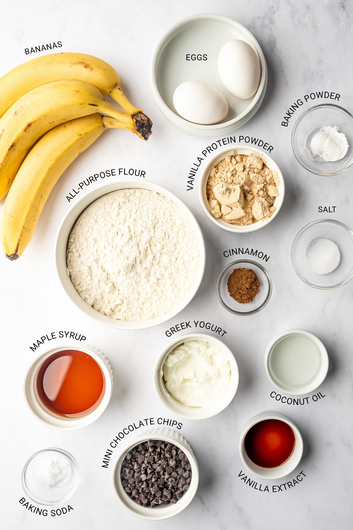Overhead layout of bananas, eggs, all-purpose flour, vanilla protein powder, baking powder, salt, cinnamon, maple syrup, Greek yogurt, coconut oil, vanilla extract, baking soda, and mini chocolate chips in white and clear bowls, with black text labels identifying each ingredient.