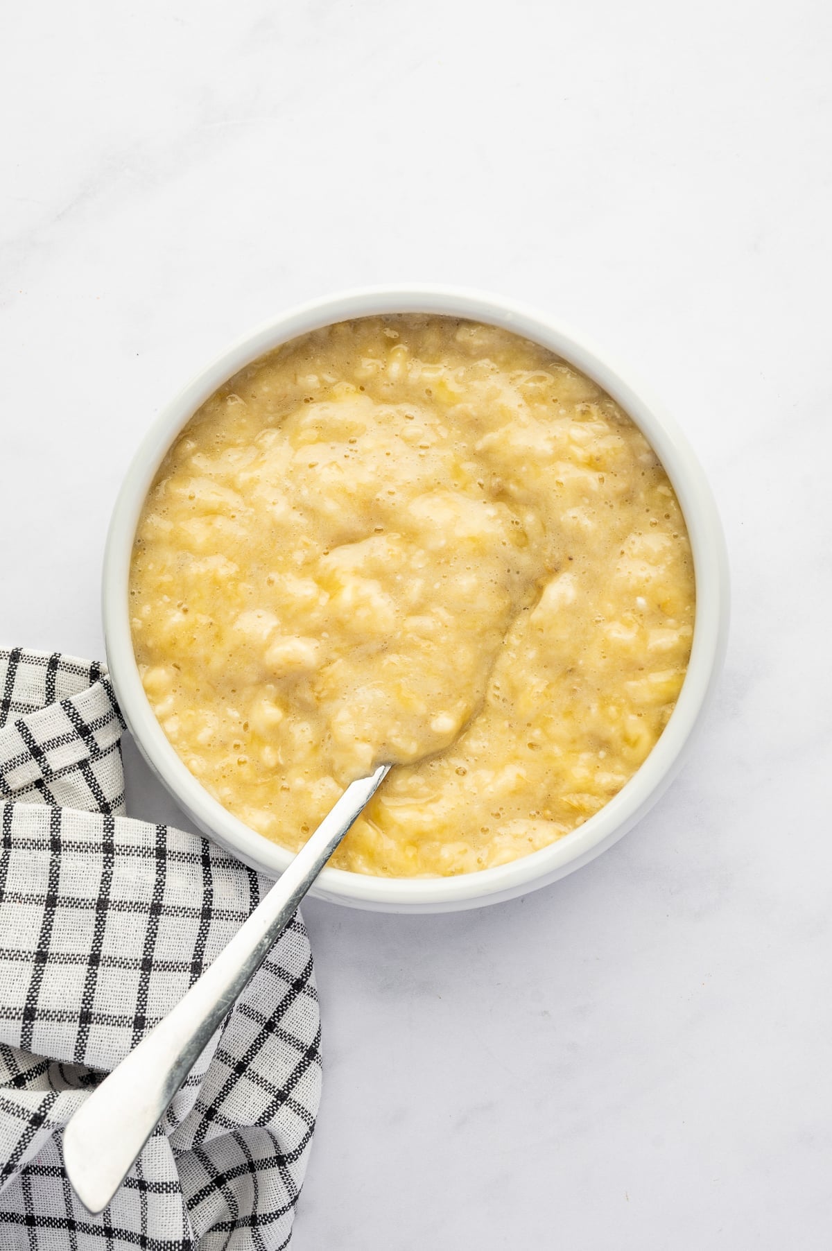 Mashed ripe bananas in a round white bowl with a silver spoon, arranged on a white marble surface beside a black-and-white checkered kitchen towel.