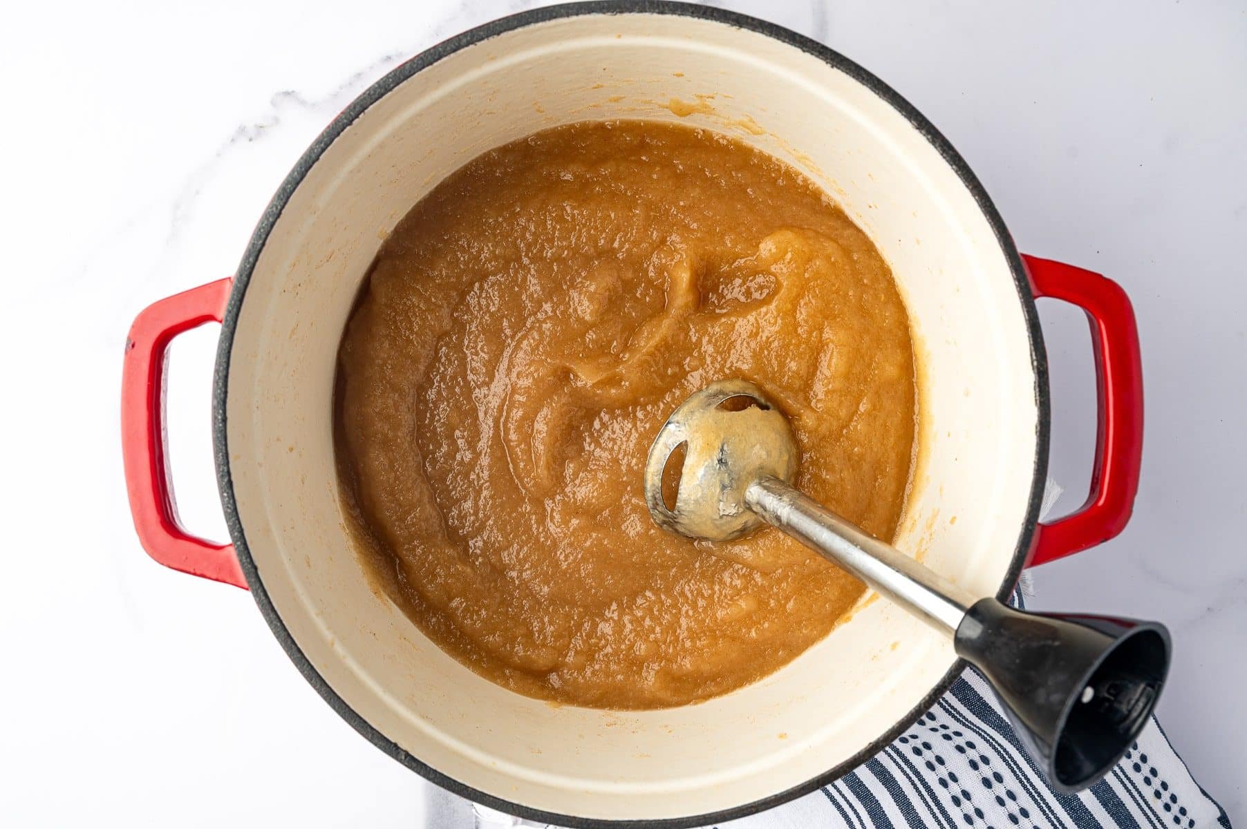 Overhead view of smooth golden-brown applesauce being blended directly in a cream-colored enameled pot with bright red handles using a stainless steel immersion blender with a black handle. A black-and-white striped towel is visible along the lower left edge.
