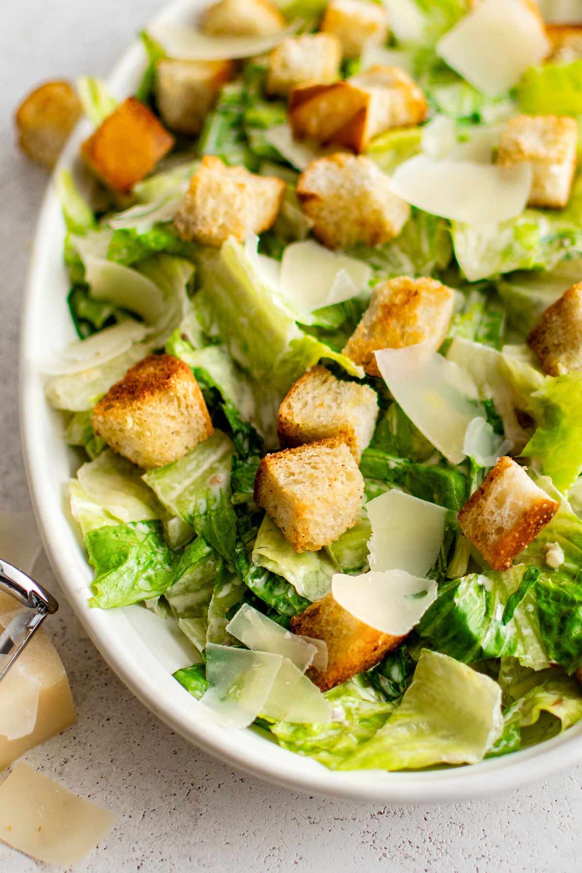 Close-up view of Caesar salad in a white oval serving platter with crisp green romaine coated in creamy dressing, golden croutons, and large pale Parmesan shavings. A metal cheese shaver and Parmesan are partially visible near the lower left edge.
