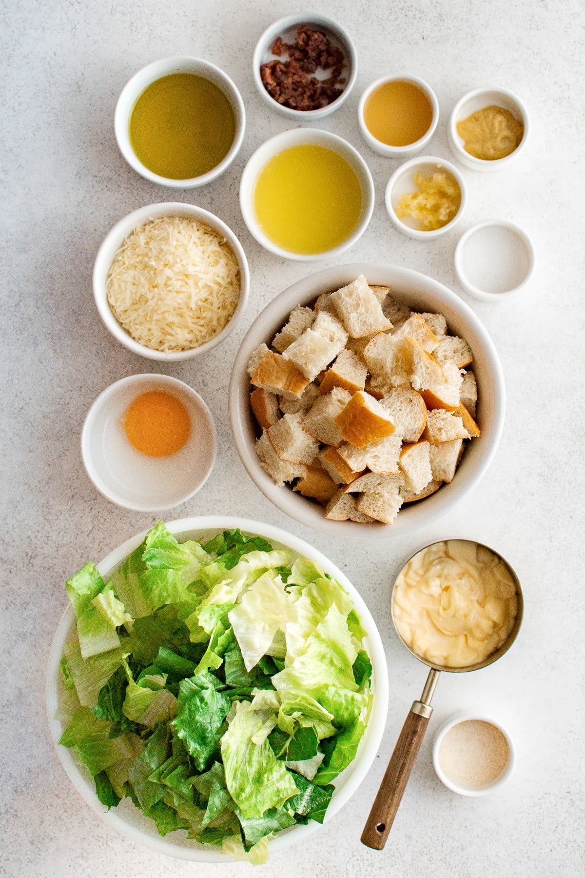 Overhead arrangement of Caesar salad ingredients on a light gray textured surface, including chopped green romaine in a large white bowl, cubed French bread in a cream bowl, mayonnaise in a metal measuring cup with a wooden handle, an egg yolk in a small white dish, grated Parmesan, olive oil, melted butter, chopped anchovies, lemon juice, stone-ground mustard, minced garlic, salt, and garlic salt in white bowls. No text overlay or garnish is present.