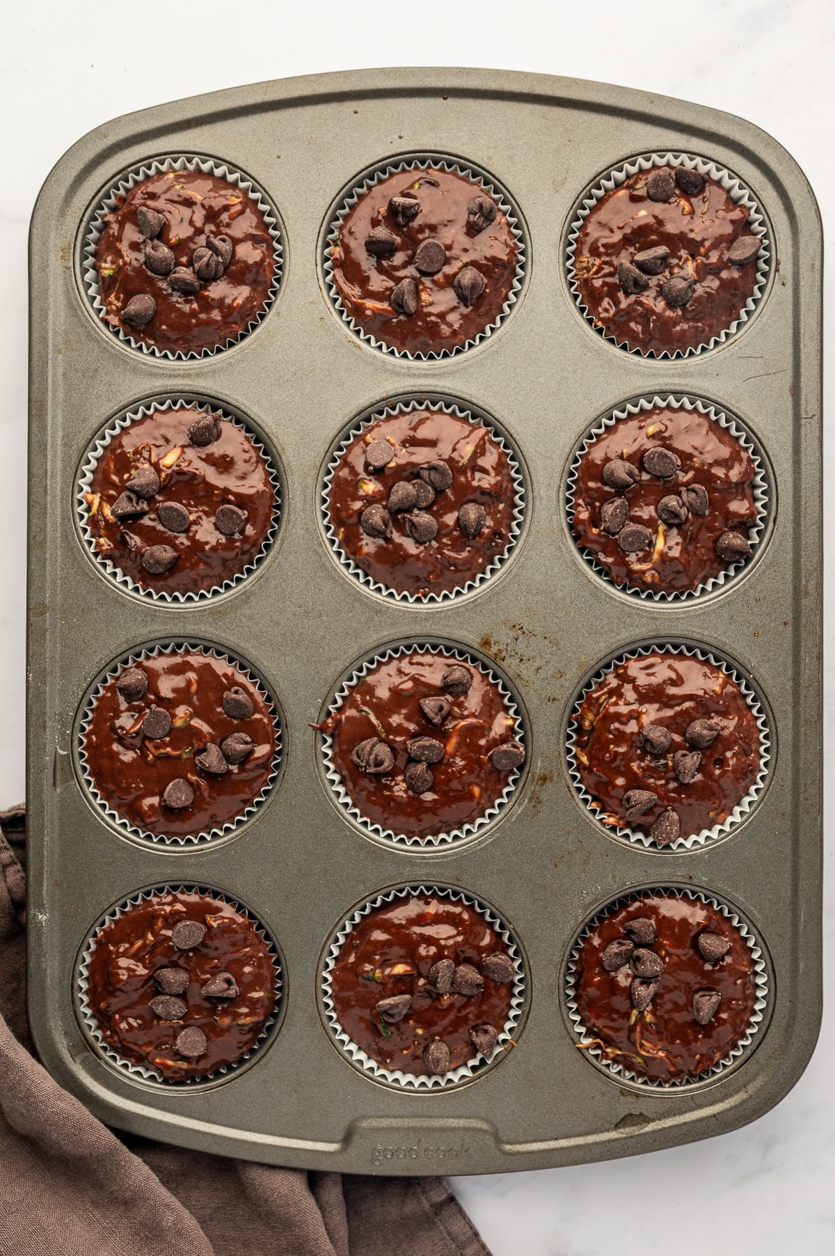 Overhead view of a gray metal 12-cup muffin pan filled with white paper liners and dark chocolate zucchini muffin batter. Each unbaked muffin is filled nearly to the top and sprinkled with semisweet chocolate chips, with small pieces of shredded zucchini visible in the batter.