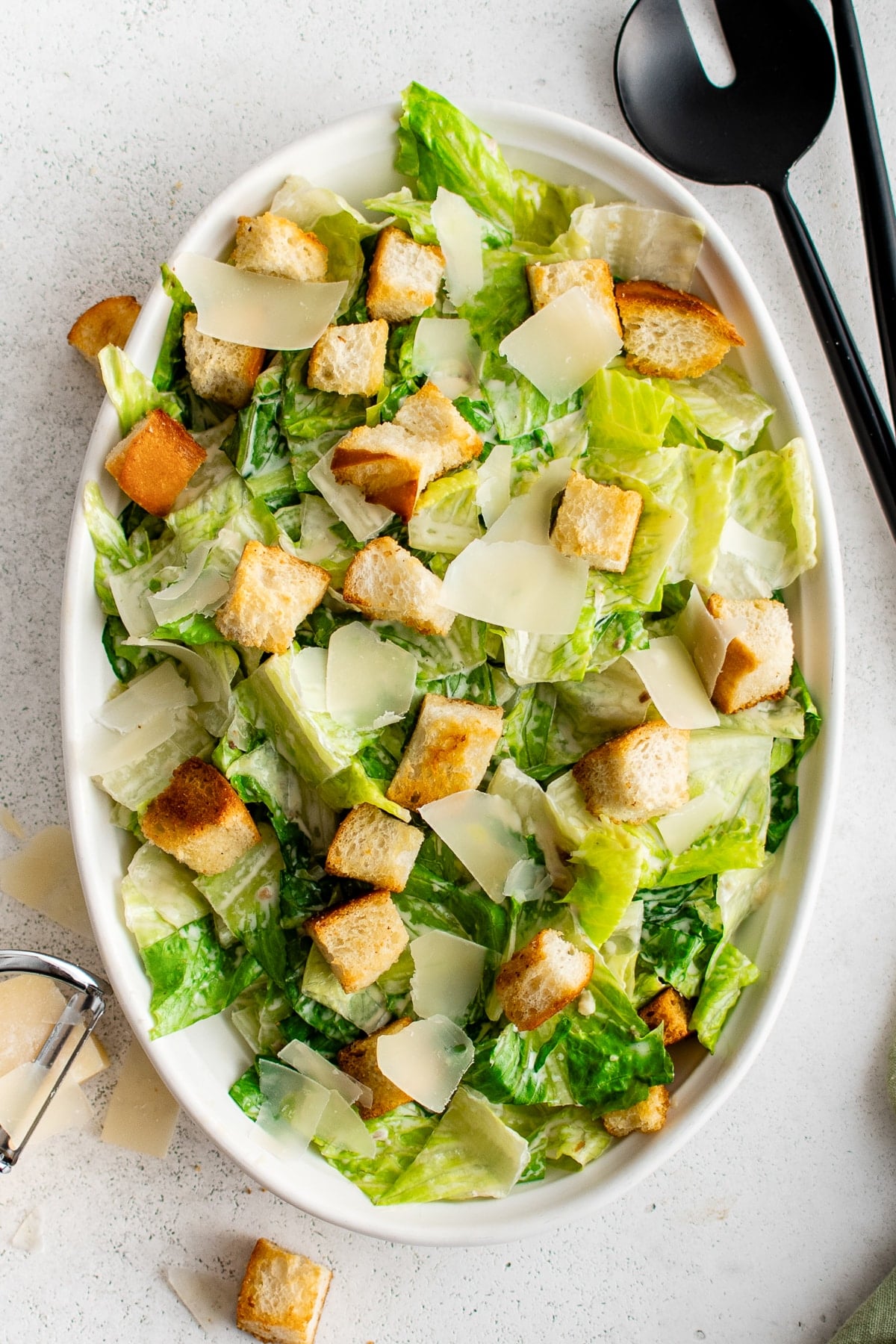 Overhead view of classic Caesar salad served in a large white oval platter with crisp green romaine, golden homemade croutons, creamy dressing, and wide shavings of Parmesan cheese. Black serving utensils sit at the upper right, while a block of Parmesan and a metal cheese shaver appear at the lower left.