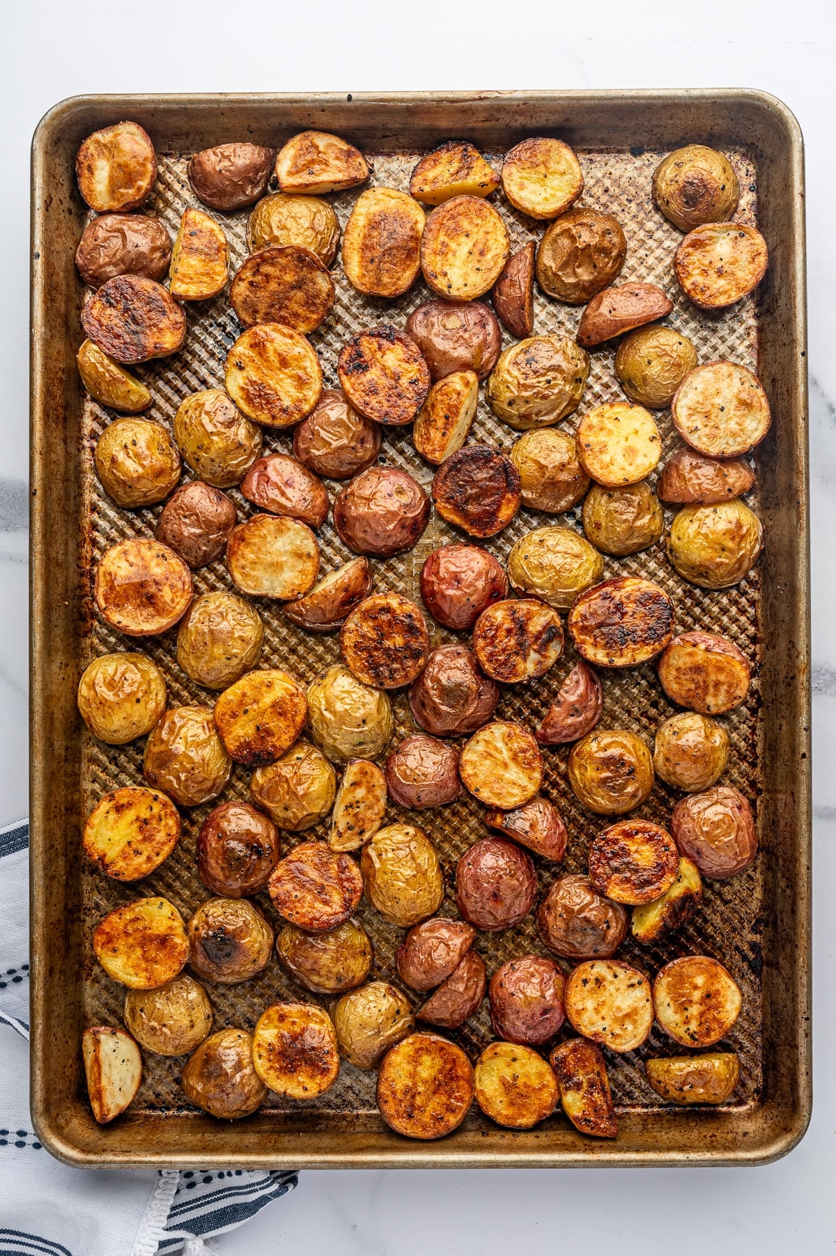 Crispy oven roasted baby red and Yukon Gold potatoes spread across a browned metal sheet pan, with deeply golden caramelized cut sides, wrinkled skins, crisp edges, and visible black pepper seasoning.