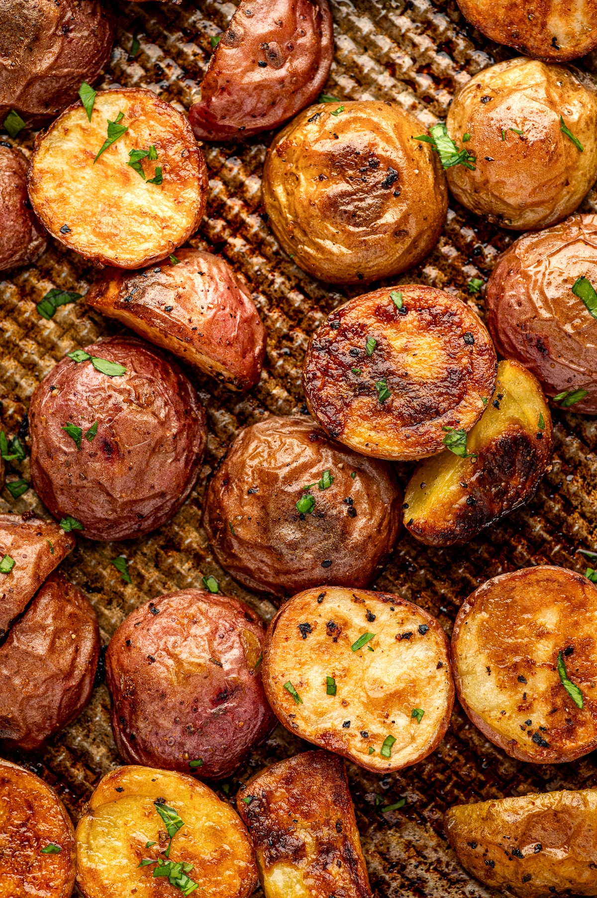 Close-up of crispy roasted red and Yukon Gold baby potatoes on a browned metal baking sheet, showing deeply caramelized golden cut sides, crisp wrinkled skins, black pepper, and a light garnish of chopped fresh parsley.