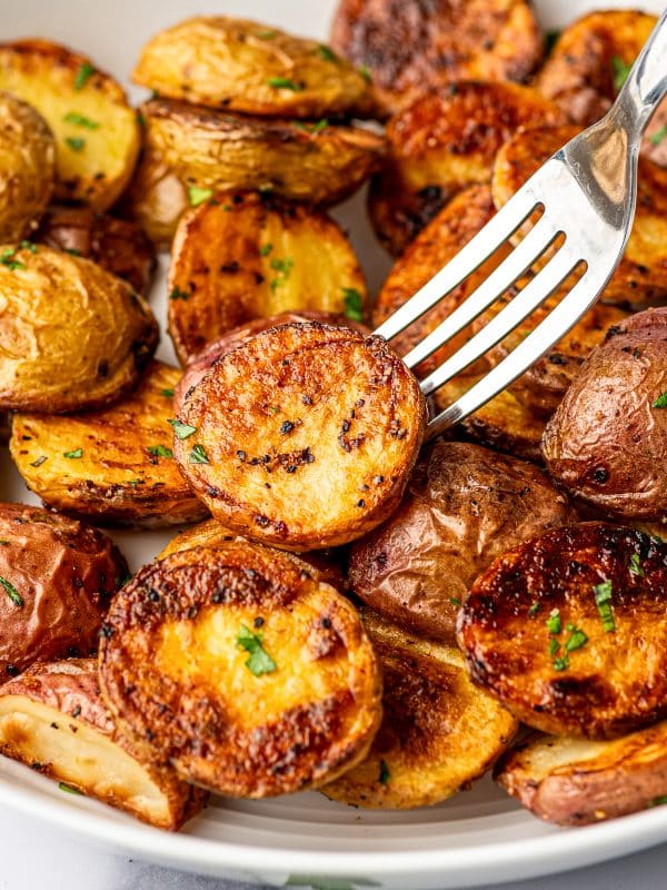 A silver fork lifting one halved roasted baby potato above a white bowl filled with crispy red and Yukon Gold potatoes, showing the potato's deeply golden caramelized cut surface, crisp browned edges, black pepper, and scattered fresh parsley.