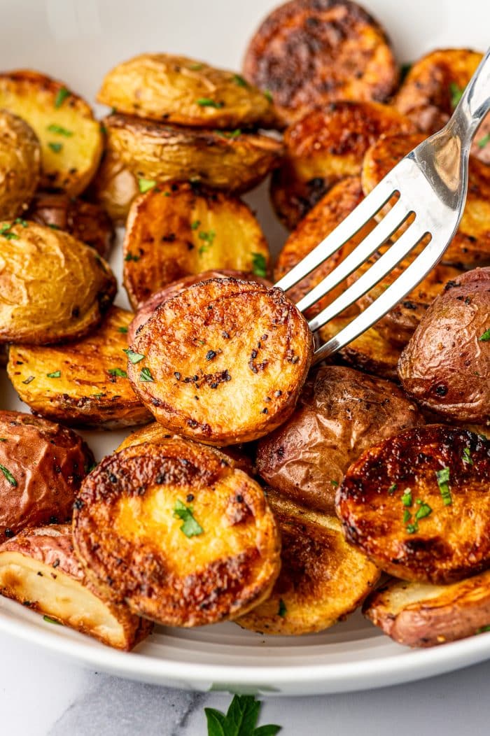 A silver fork lifting one halved roasted baby potato above a white bowl filled with crispy red and Yukon Gold potatoes, showing the potato's deeply golden caramelized cut surface, crisp browned edges, black pepper, and scattered fresh parsley.