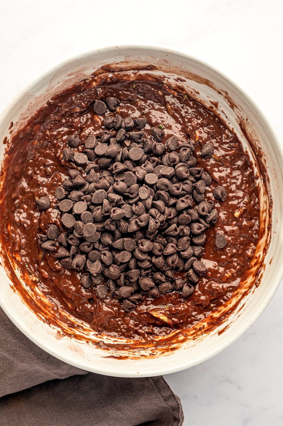 Overhead view of thick dark chocolate zucchini muffin batter in an off-white ceramic bowl topped with a generous mound of semisweet chocolate chips. Small green flecks of shredded zucchini are visible throughout the glossy chocolate batter.