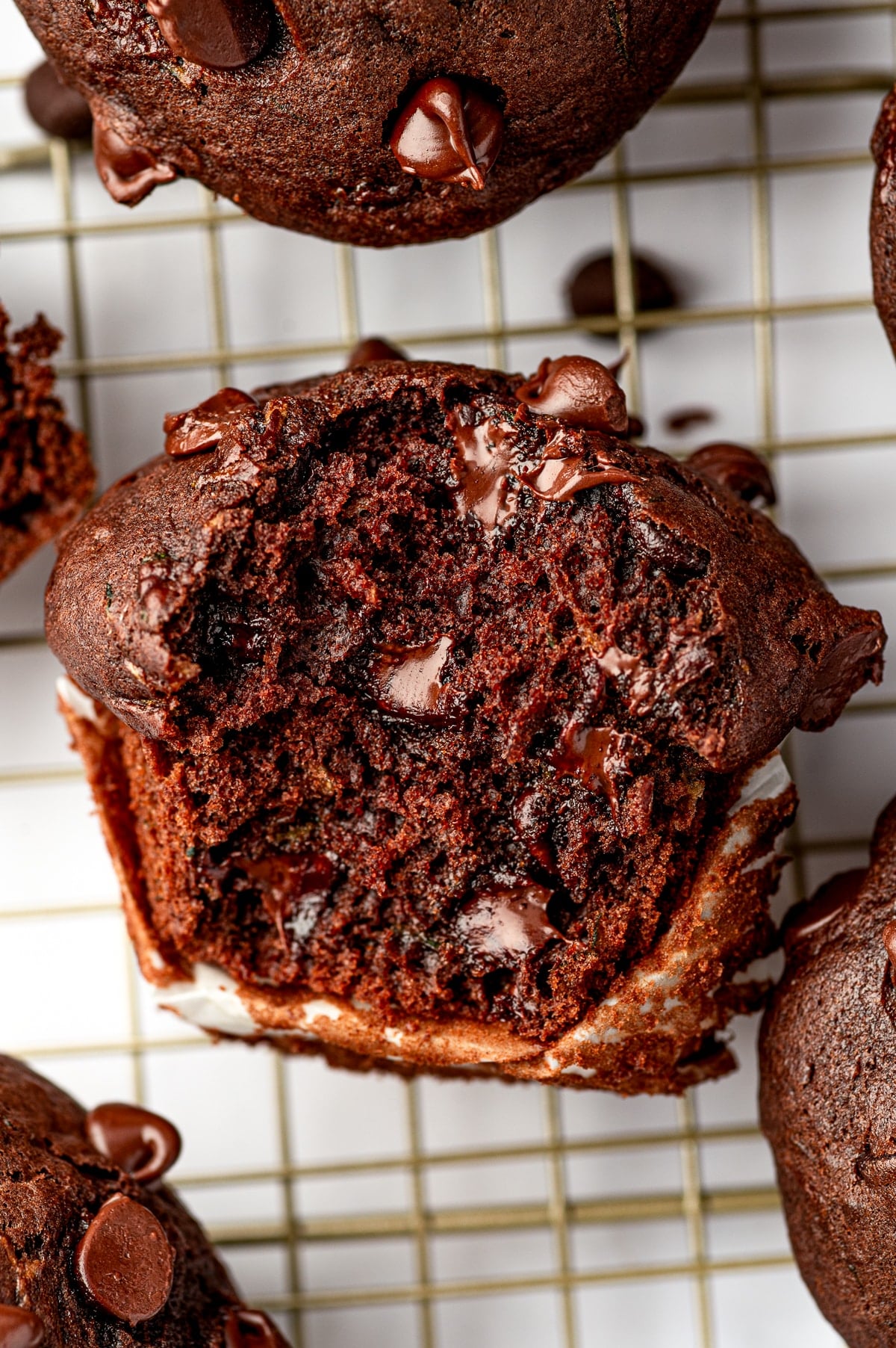 Close-up of a double chocolate zucchini muffin torn open on a gold wire cooling rack, exposing its dark, tender crumb and several pockets of soft melted chocolate. Whole chocolate zucchini muffins topped with chocolate chips surround the open muffin.
