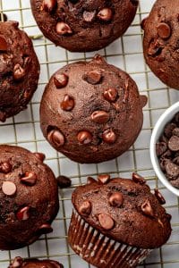 Overhead view of several double chocolate zucchini muffins cooling on a gold wire rack over a white surface. The dark chocolate muffins have domed tops covered with semisweet chocolate chips, and a small white bowl filled with additional chocolate chips sits along the right edge.