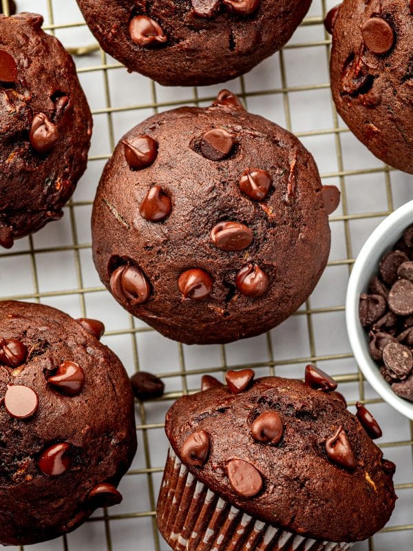 Overhead view of several double chocolate zucchini muffins cooling on a gold wire rack over a white surface. The dark chocolate muffins have domed tops covered with semisweet chocolate chips, and a small white bowl filled with additional chocolate chips sits along the right edge.
