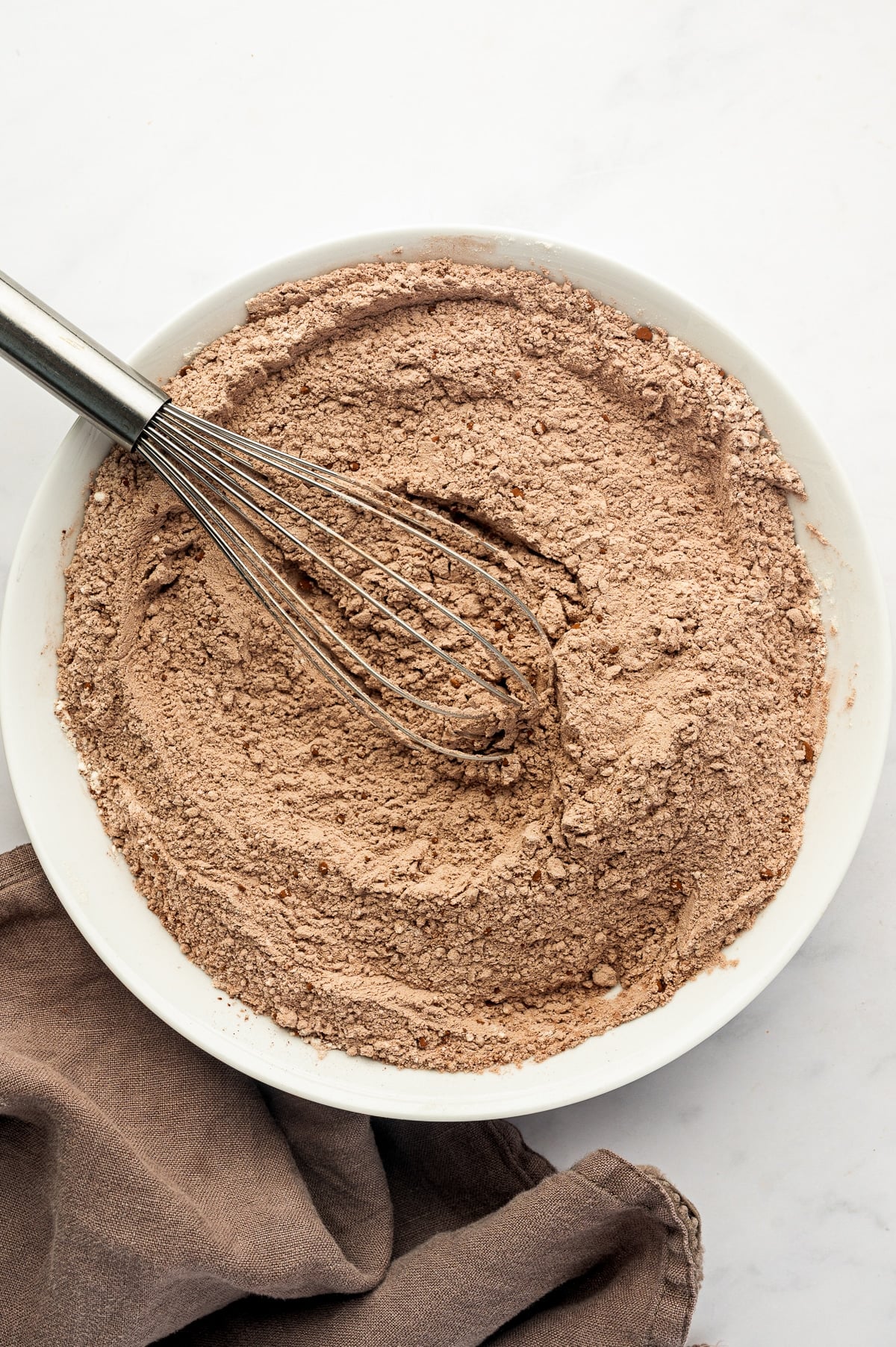Overhead view of the dry ingredients for double chocolate zucchini muffins whisked together in a large white ceramic bowl, with cocoa powder giving the flour mixture a light brown color. A stainless steel whisk rests inside the bowl, with a brown cloth napkin along the lower left edge.