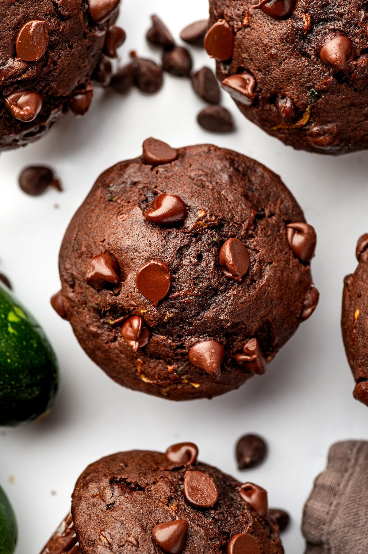 Overhead arrangement of double chocolate zucchini muffins on a white surface, centered around one large dark chocolate muffin topped with glossy chocolate chips. Additional muffins and scattered chocolate chips surround it, with a whole green zucchini along the lower left and a brown cloth napkin at the lower right.