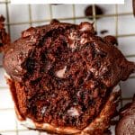 Vertical Pinterest graphic featuring a close-up of a double chocolate zucchini muffin torn open to reveal a dark, tender crumb with pockets of melted chocolate chips. A white text box at the top reads "Double Chocolate Zucchini Muffins" in black lettering with a pale pink underline, and "THEFORKEDSPOON.COM" appears in black at the bottom.