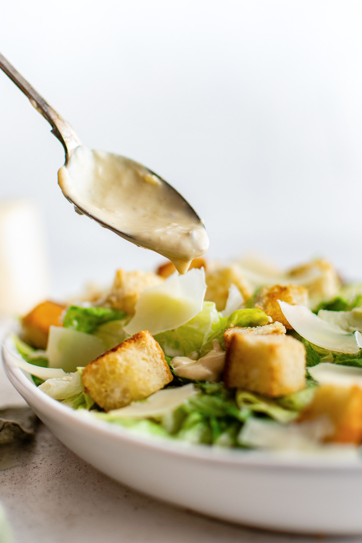 Close-up side view of a silver spoon dripping thick homemade Caesar dressing over a white bowl filled with green romaine lettuce, golden croutons, and shaved Parmesan cheese. The salad and background are softly blurred, and no text overlay is present.