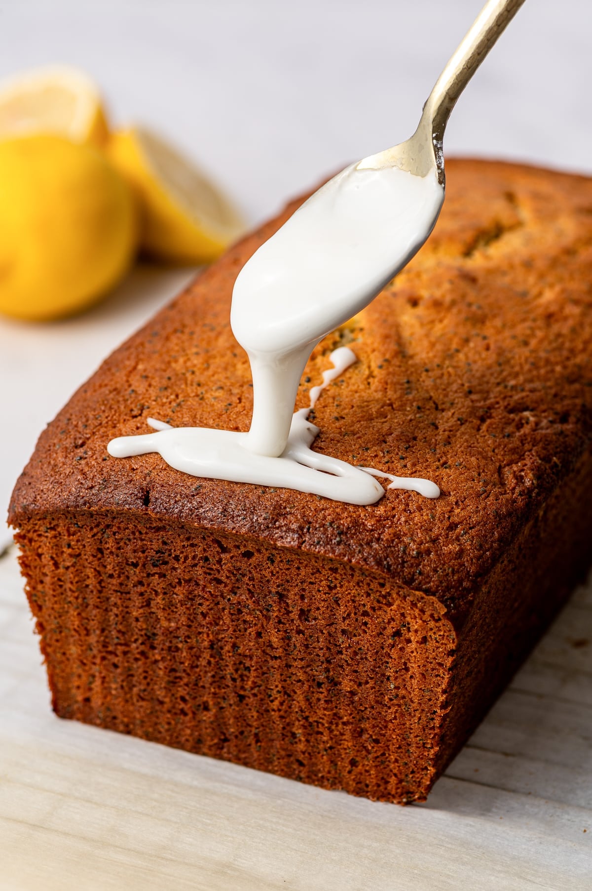 Close three-quarter view of a deeply golden lemon poppy seed loaf on a light wooden board as thick white lemon glaze drips from a gold spoon onto the top. Whole and halved yellow lemons appear softly blurred in the background.