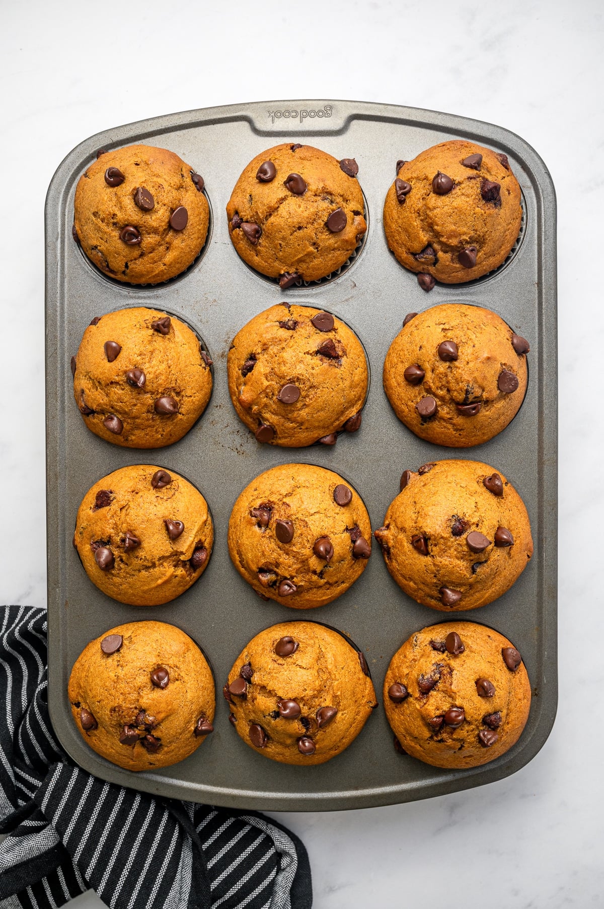 Twelve freshly baked pumpkin chocolate chip muffins with tall golden-orange domed tops and semi-sweet chocolate chips in a gray metal muffin pan, photographed overhead on a white marble surface with a black-and-white striped towel.