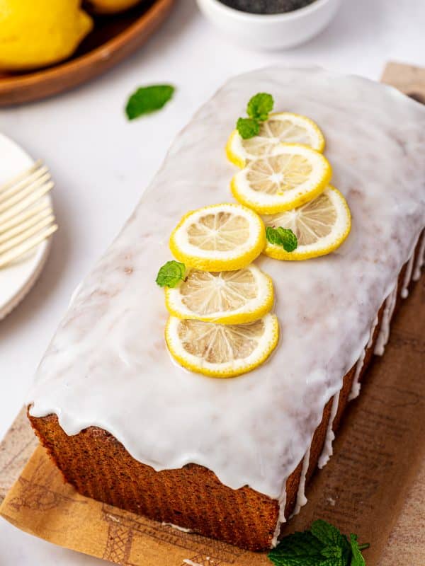 A whole golden-brown lemon poppy seed loaf covered with thick white lemon glaze and arranged on a brown printed serving board. Thin yellow lemon slices and small fresh mint leaves garnish the top, with whole lemons, a white bowl of black poppy seeds, white plates, and a gold fork in the background.