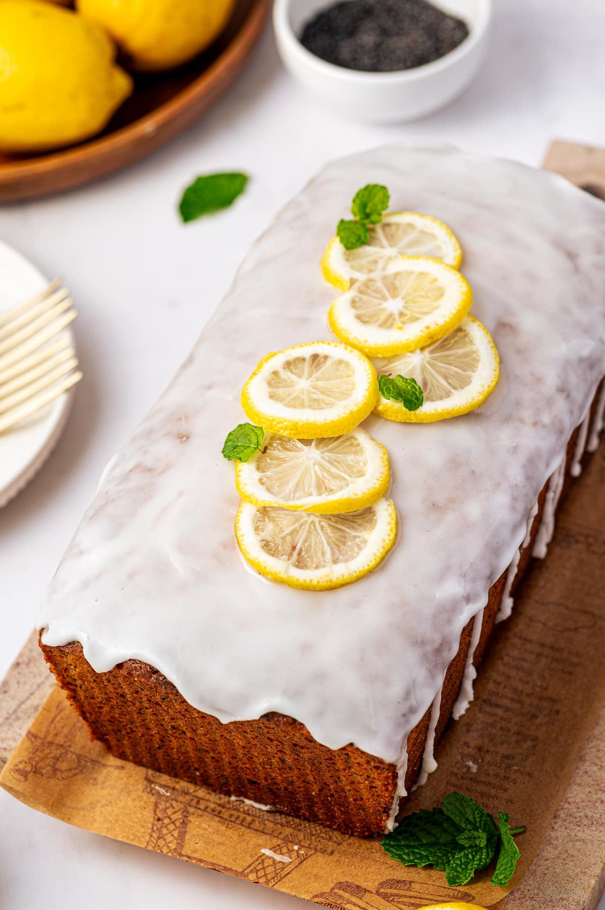 A whole golden-brown lemon poppy seed loaf covered with thick white lemon glaze and arranged on a brown printed serving board. Thin yellow lemon slices and small fresh mint leaves garnish the top, with whole lemons, a white bowl of black poppy seeds, white plates, and a gold fork in the background.