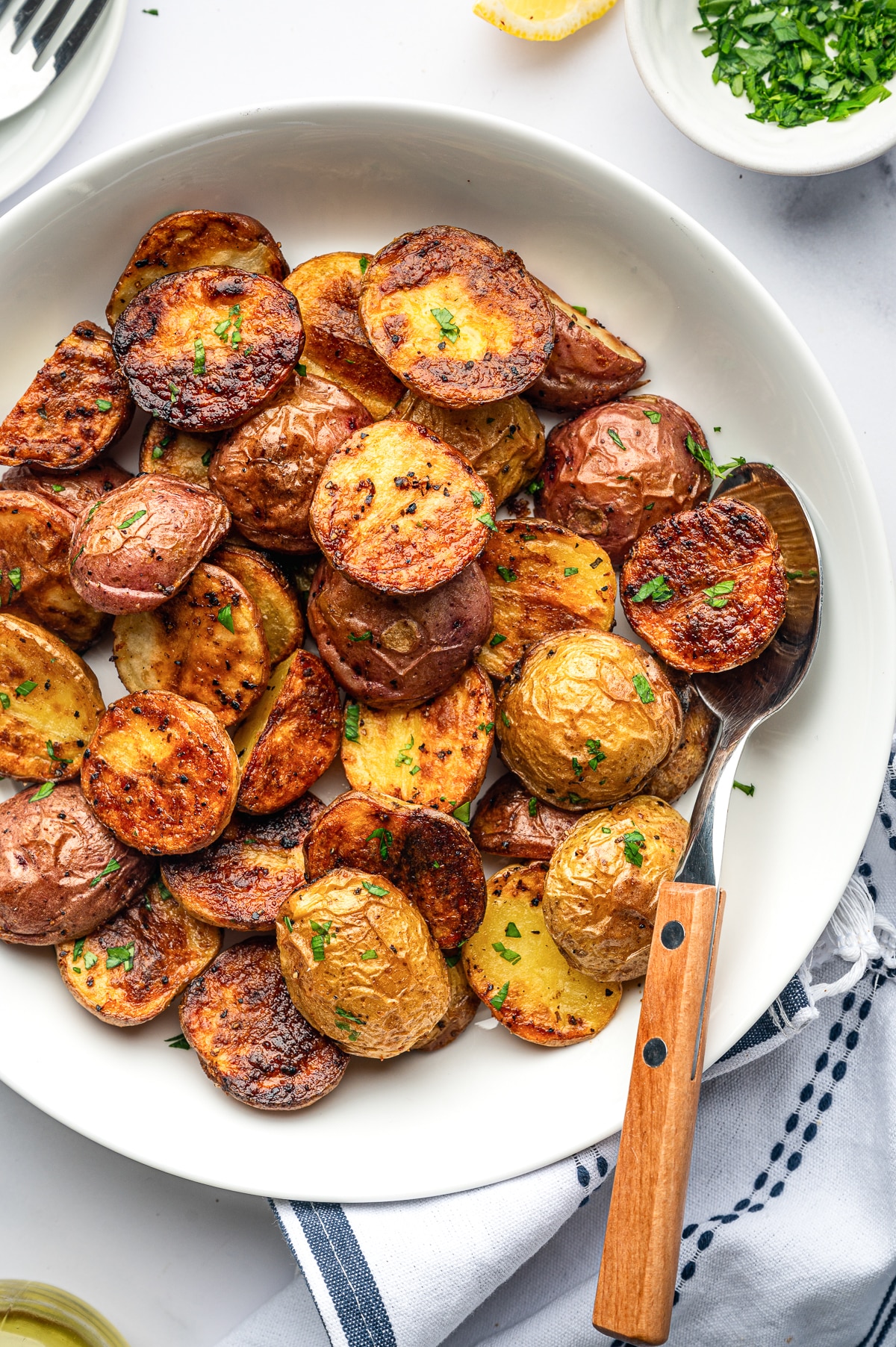Close overhead view of golden roasted red and Yukon Gold baby potatoes in a large white bowl, showing deeply browned cut sides and crisp skins with chopped parsley, a metal serving spoon with a wooden handle, a lemon wedge, chopped parsley in a small white bowl, and a blue-and-white striped towel.
